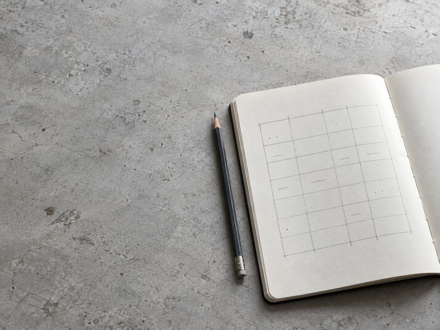An open notebook with a pencil-ruled grid and faint marks, a pencil beside it on a grey surface.