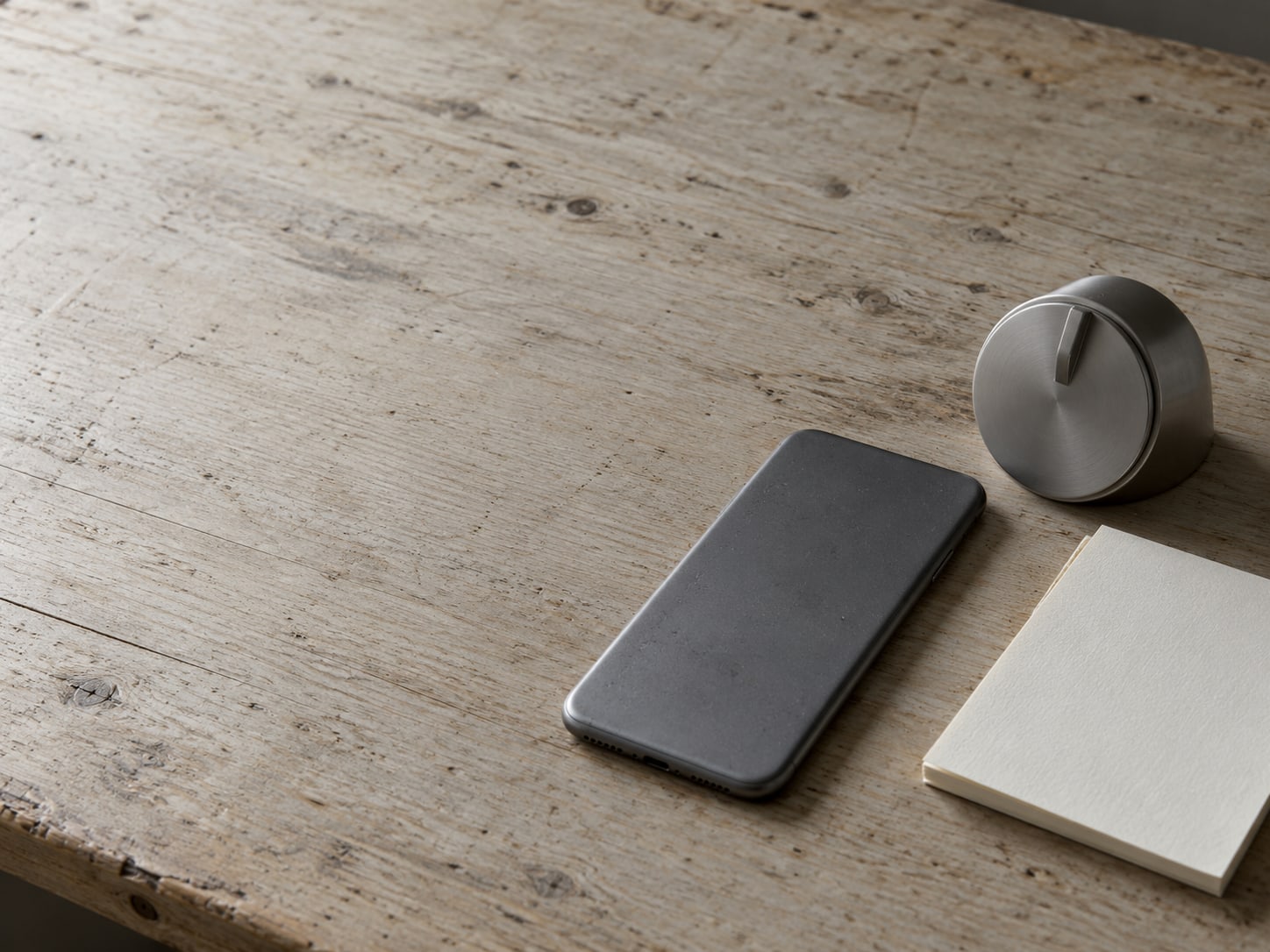 A smartphone lying face down on a desk next to a plain kitchen timer and a blank notepad.
