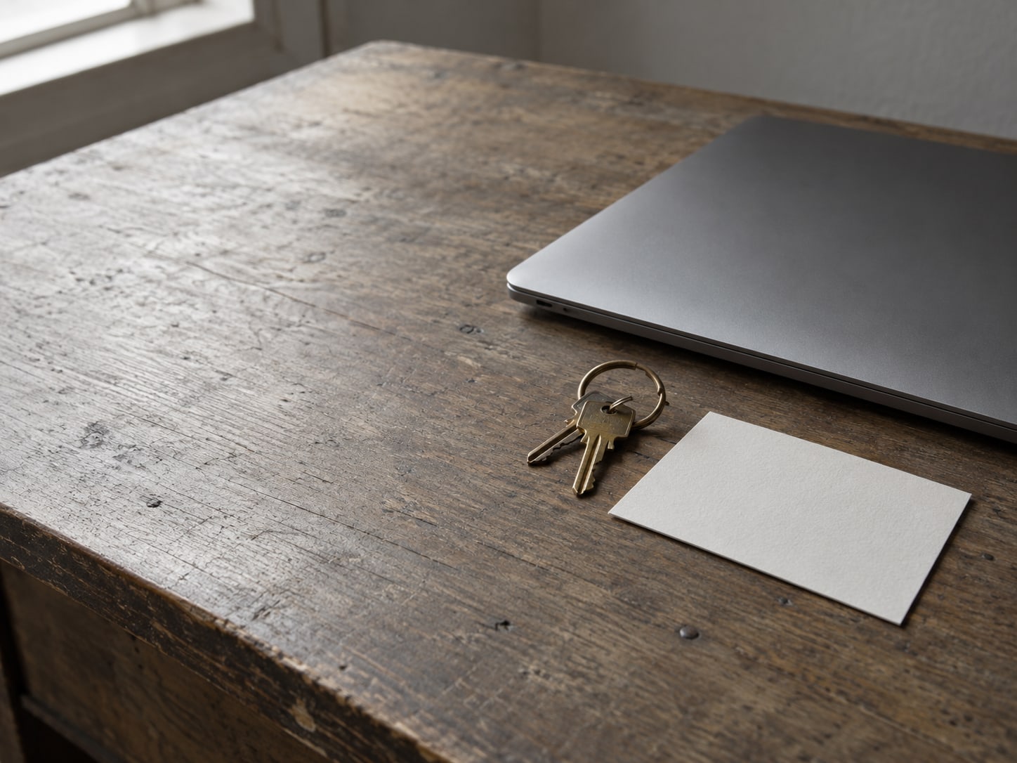 A closed laptop beside a small brass key ring and a blank index card on a worn wooden desk.
