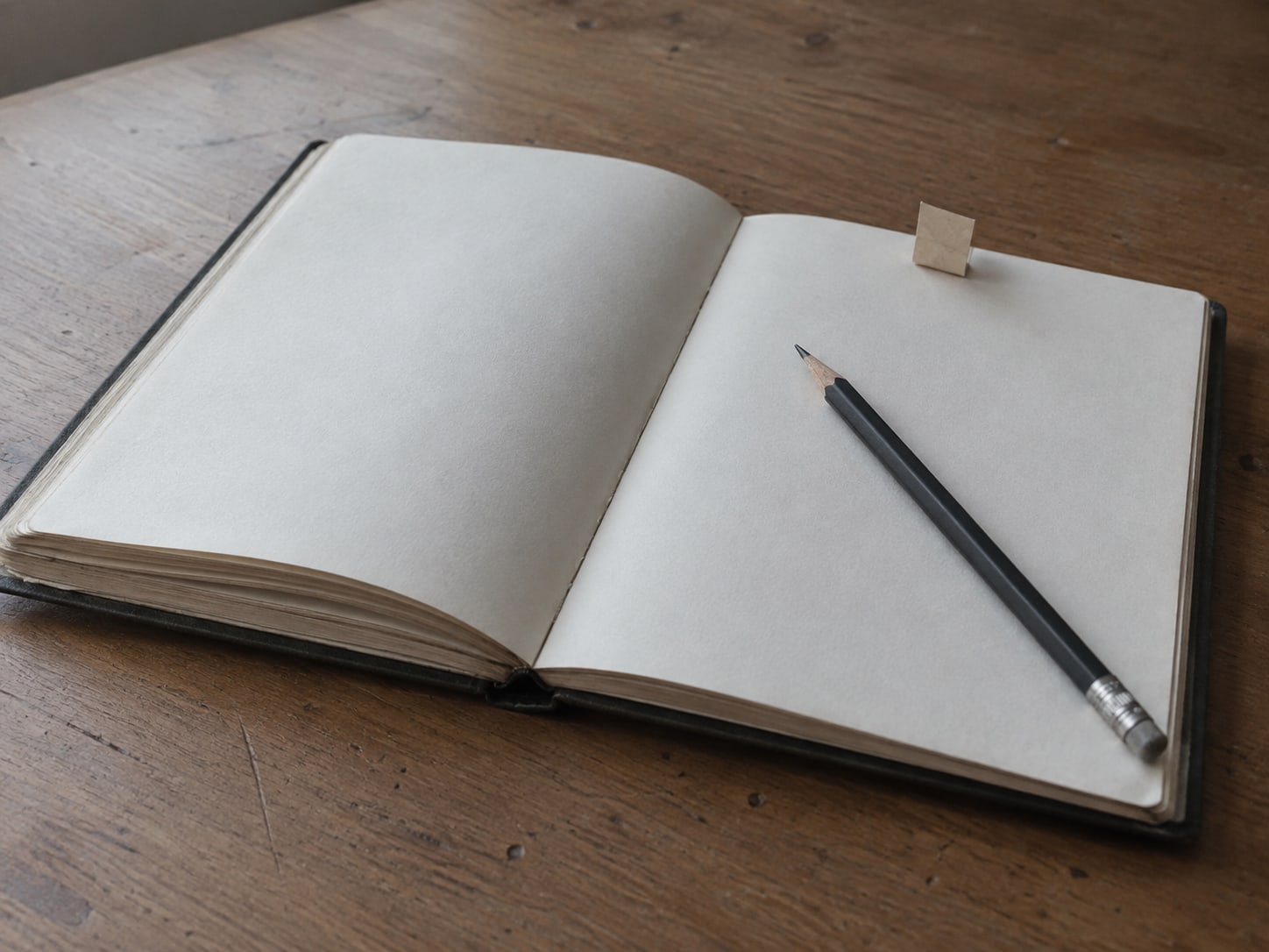 A desk planner open to blank pages with a pencil and a folded sticky tab marking one page, on a plain wooden desk.