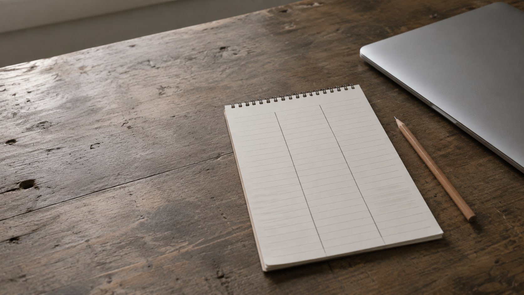 A blank ruled notepad with three pencil-drawn columns and a sharpened pencil beside a closed laptop on a wooden desk.