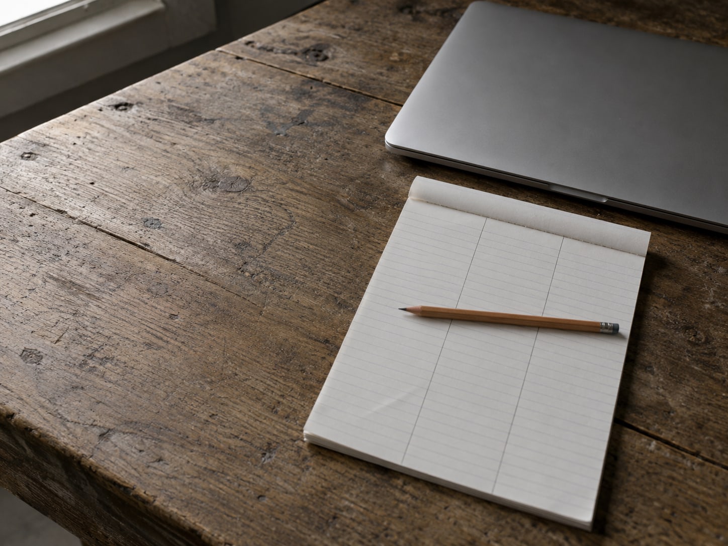 A blank ruled notepad ruled into three columns with a pencil resting across it, beside a closed laptop on a wooden desk.