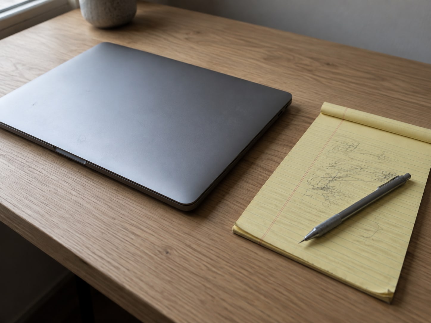 Una laptop cerrada junto a una libreta amarilla con marcas de lápiz ilegibles y un lapicero mecánico sobre un escritorio de roble claro.