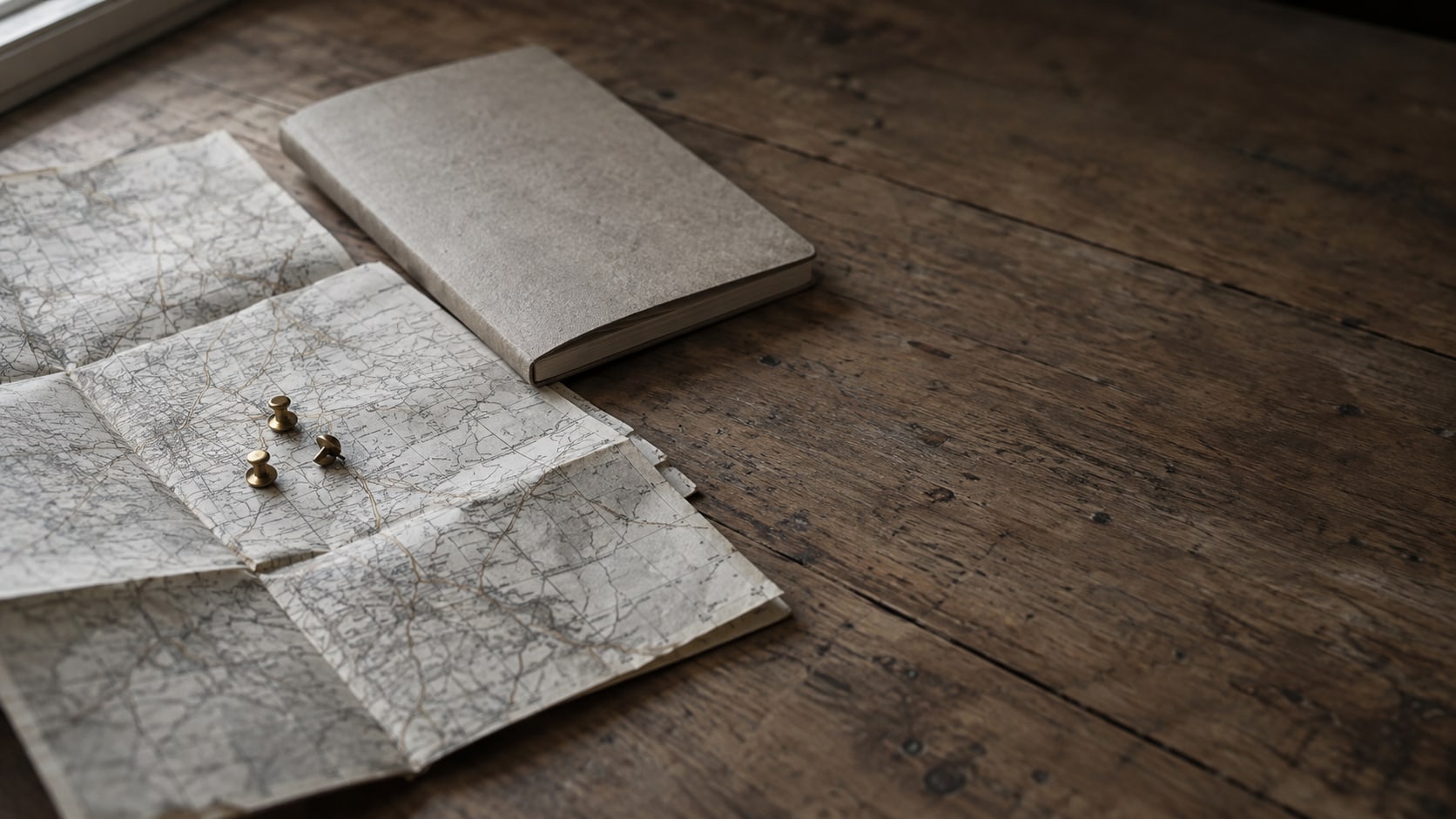 A folded paper road map on a wooden table with three brass pushpins clustered near the center and a closed notebook beside it.