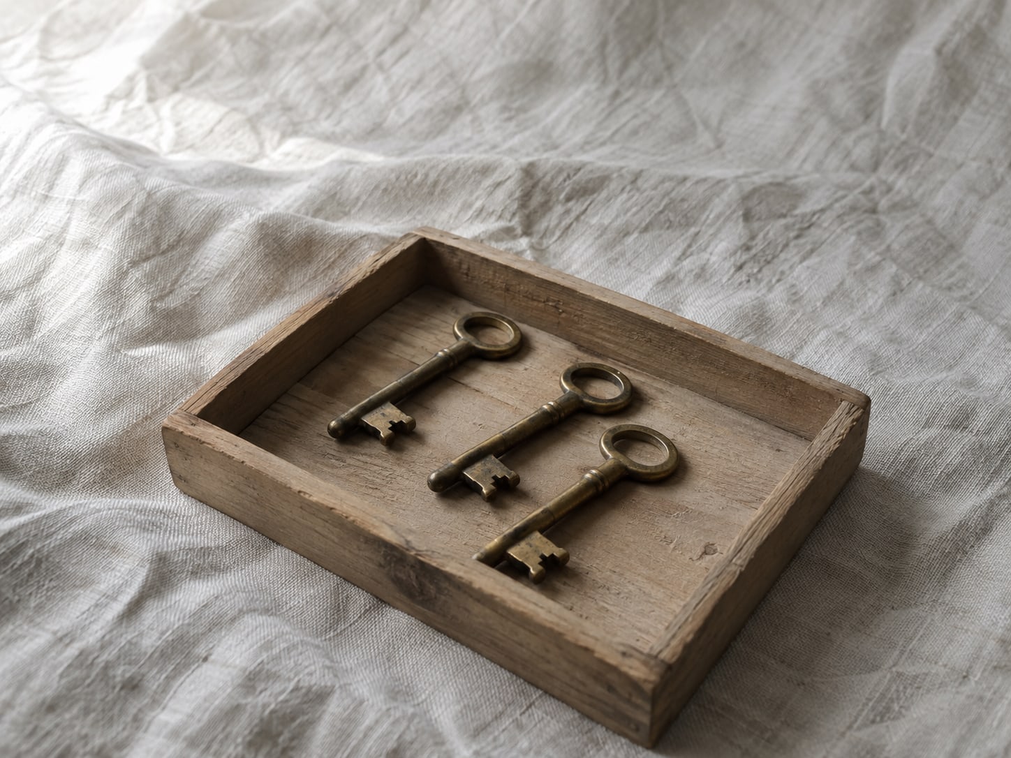 A small wooden tray holding three identical brass keys, one set slightly forward of the others, on a pale linen cloth.