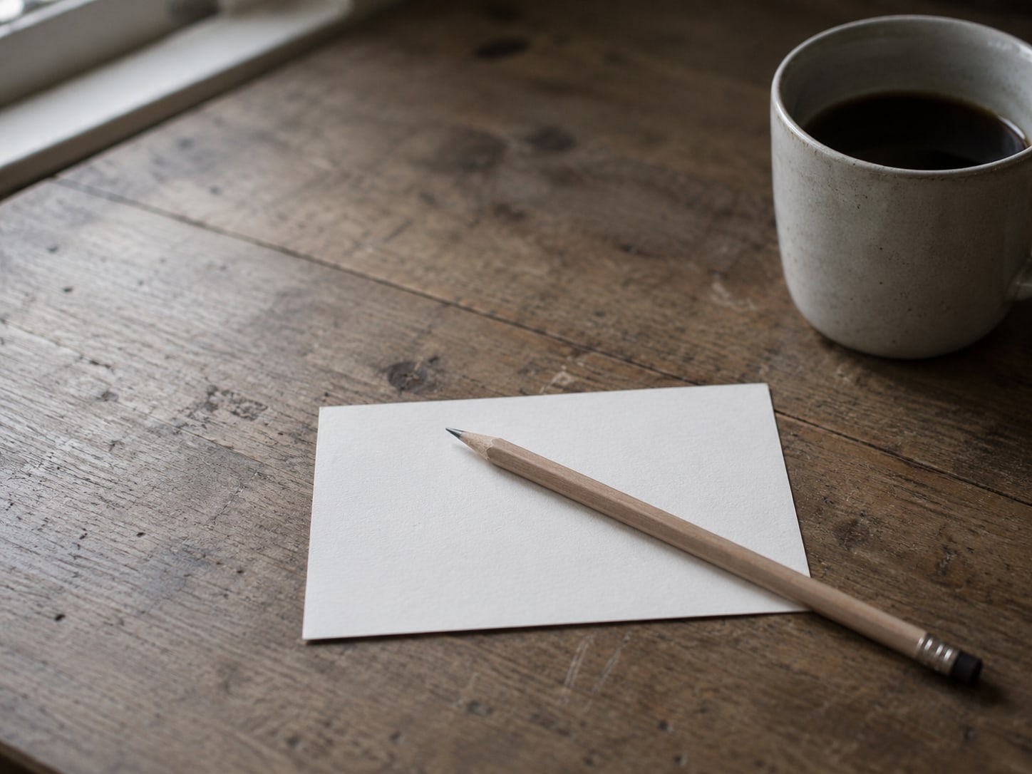 A single blank index card on a worn desk, a sharpened pencil lying across it, a coffee cup set to one side.