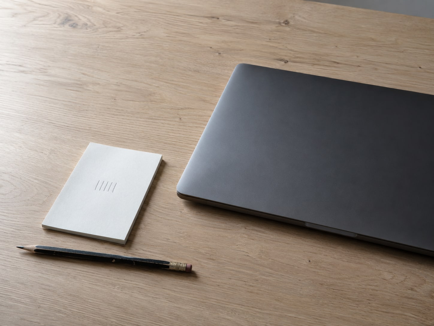 A closed laptop, a small notepad with a pencil tally of short vertical marks, and a flat pencil on a clean desk.