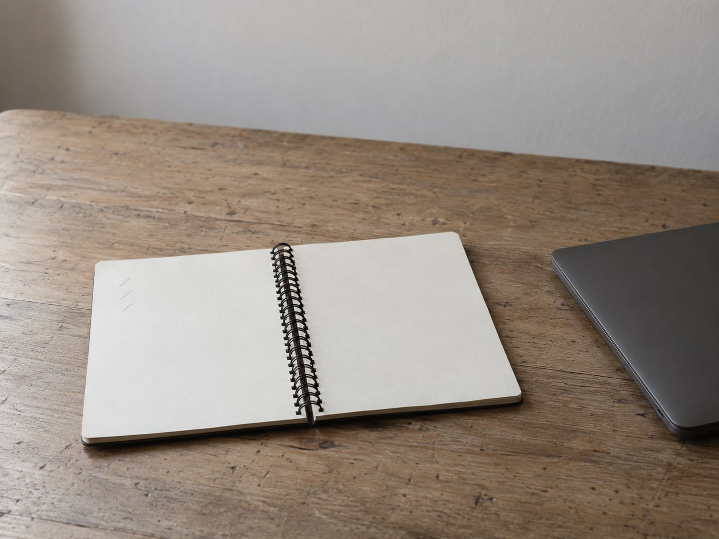 A small spiral notebook lying open on a wooden desk, pages blank except for a few pencil lines, beside a closed laptop.