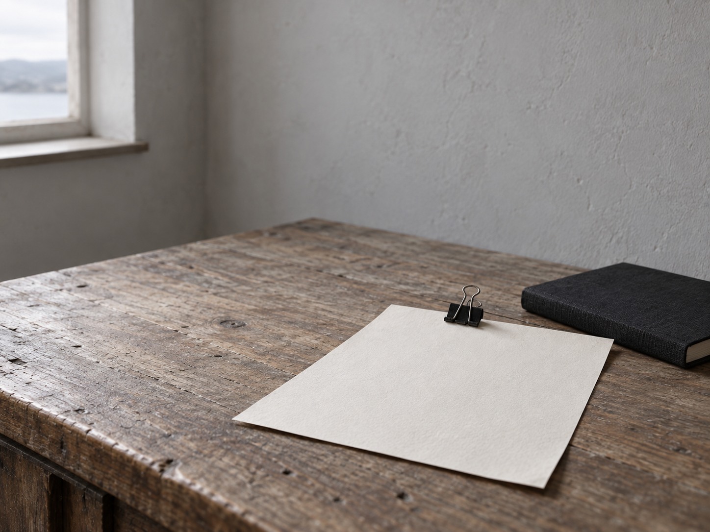 A single sheet of blank paper held by a black binder clip on a wooden desk next to a closed notebook.