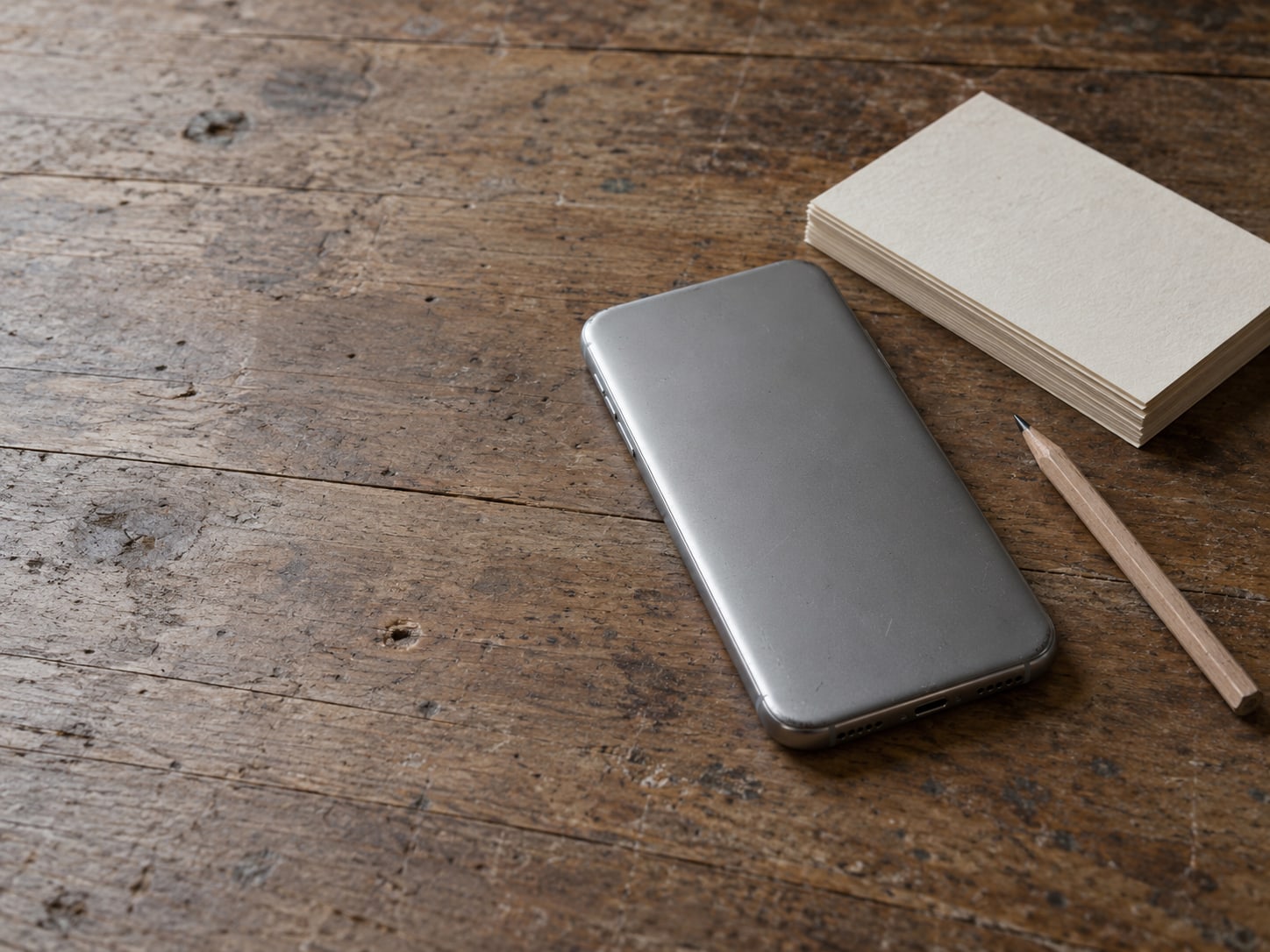 A closed smartphone lying face down on a wooden desk beside a stack of blank index cards and a pencil.