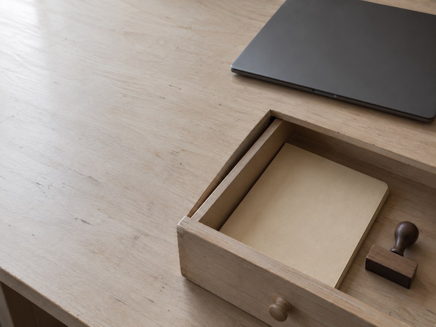 A single wooden desk drawer pulled open with a sealed folder and a rubber stamp inside, beside a closed laptop.