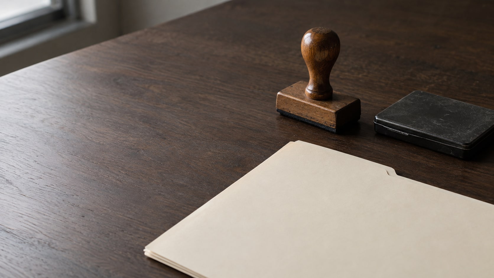 A wooden rubber stamp and a closed ink pad beside a blank cream folder on a dark walnut desk.