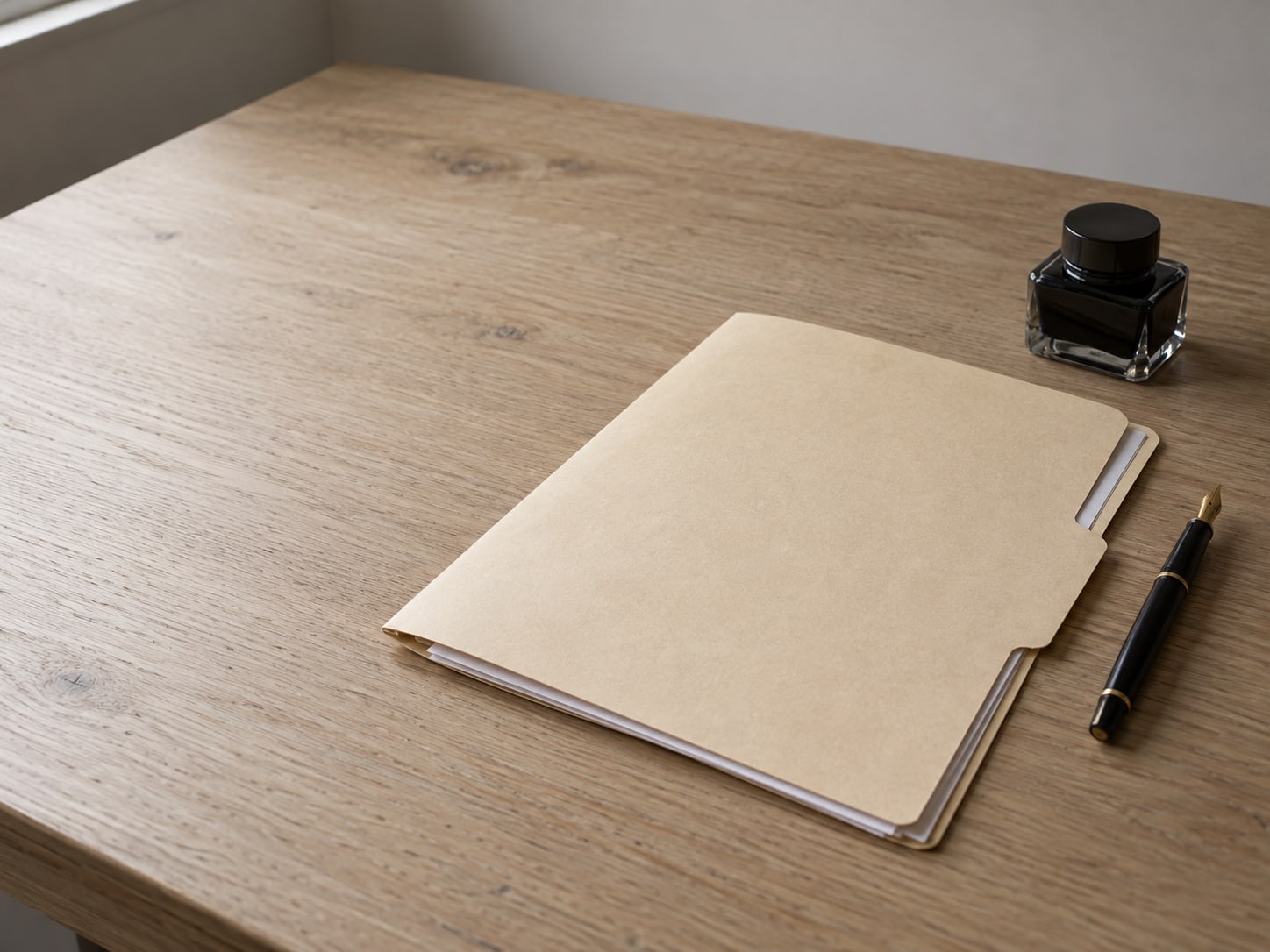 A plain manila folder holding a few blank sheets beside a fountain pen and a closed ink bottle on a light oak desk.