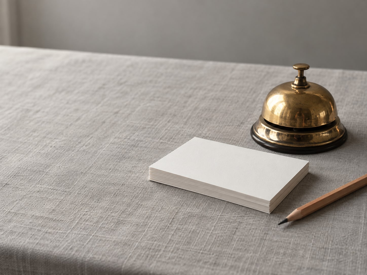 A brass desk bell beside a neat stack of blank index cards and a sharpened pencil on a grey linen tablecloth.