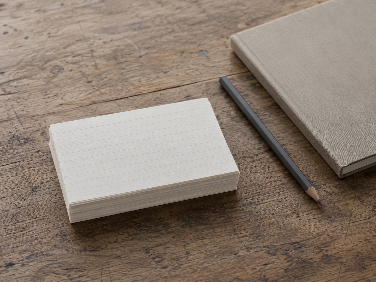 A short stack of index cards with blank lines on a worn wooden desk, beside a closed notebook and a pencil.