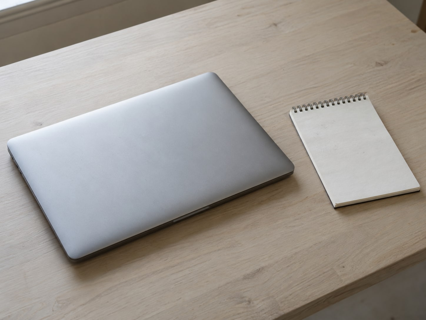 A closed laptop beside a small notepad with faint pencil marks on a clean desk.