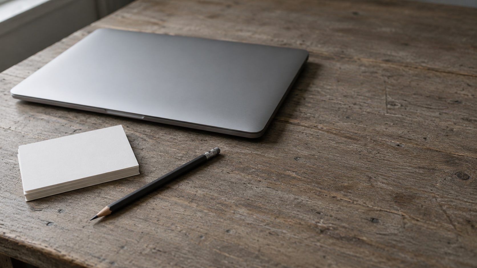 A closed laptop and a short stack of blank index cards on a worn wooden desk beside a pencil.