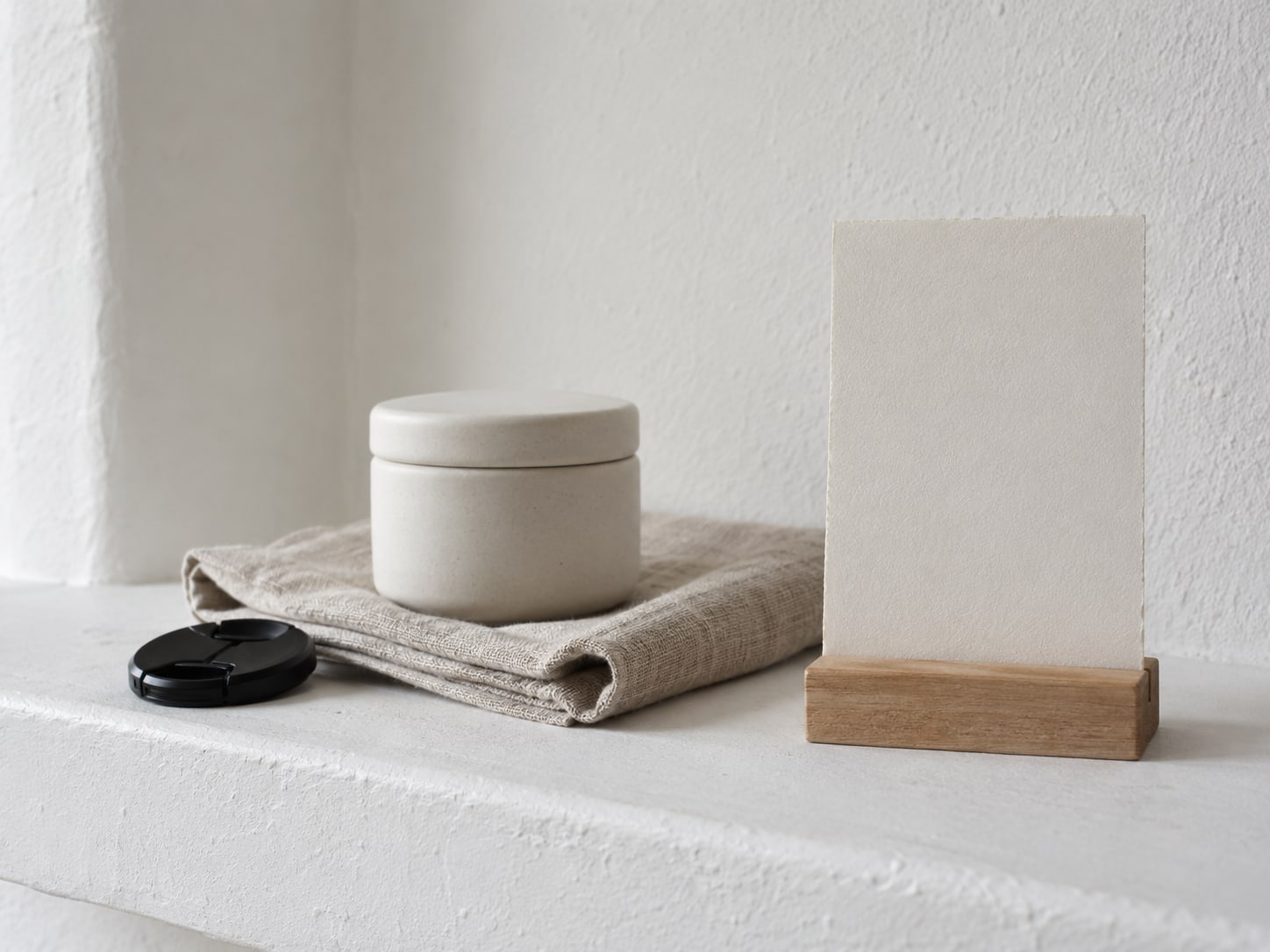 A camera lens cap and a small product sample set on a folded cloth, next to a blank card stand on a plain white shelf.