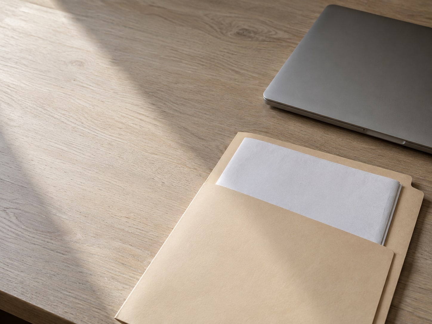 A folded sheet of blank printer paper tucked into a manila folder beside a closed laptop on a pale oak desk.
