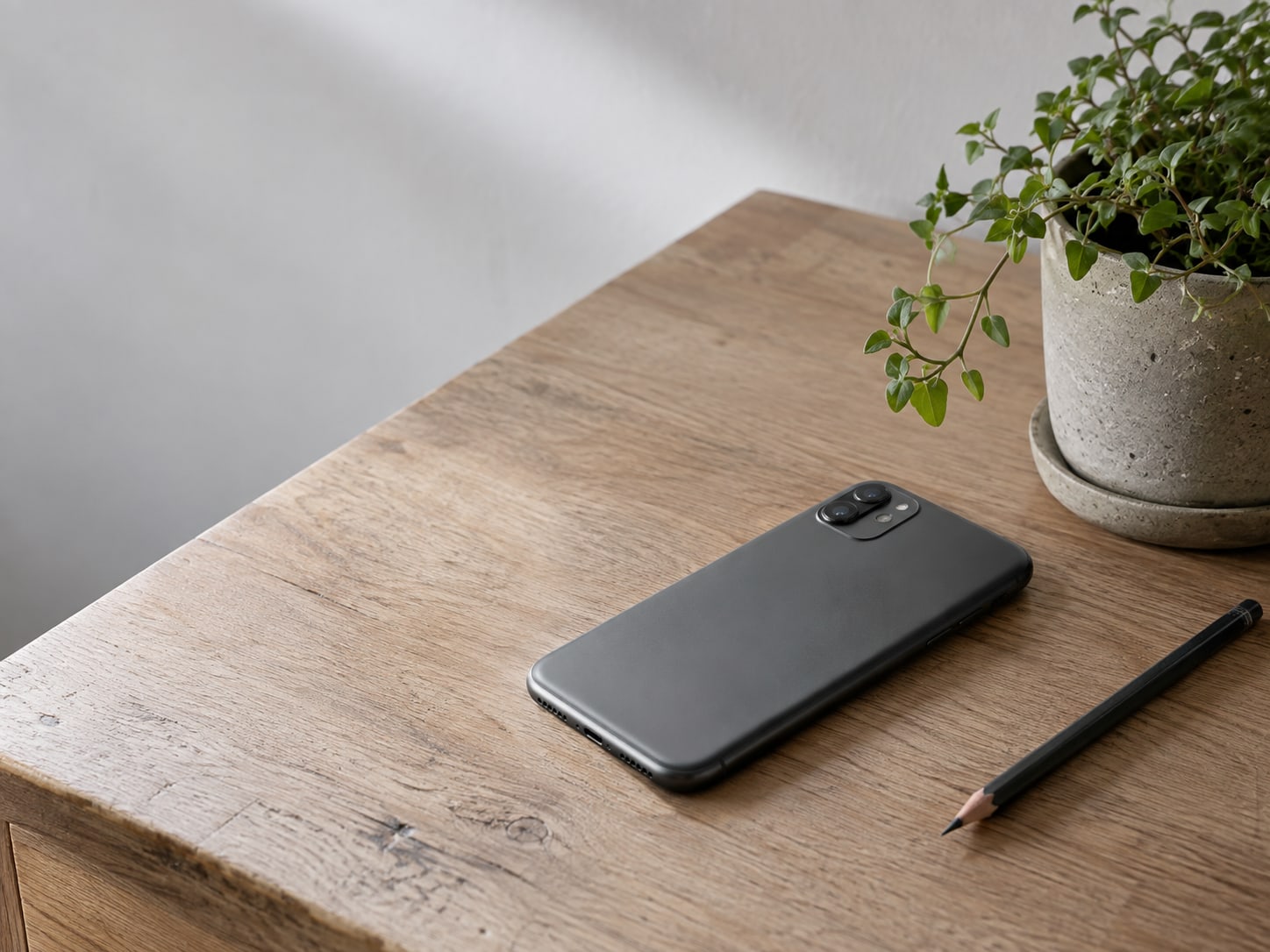 A closed smartphone lying face down on a wooden counter beside a small potted plant and a pencil.