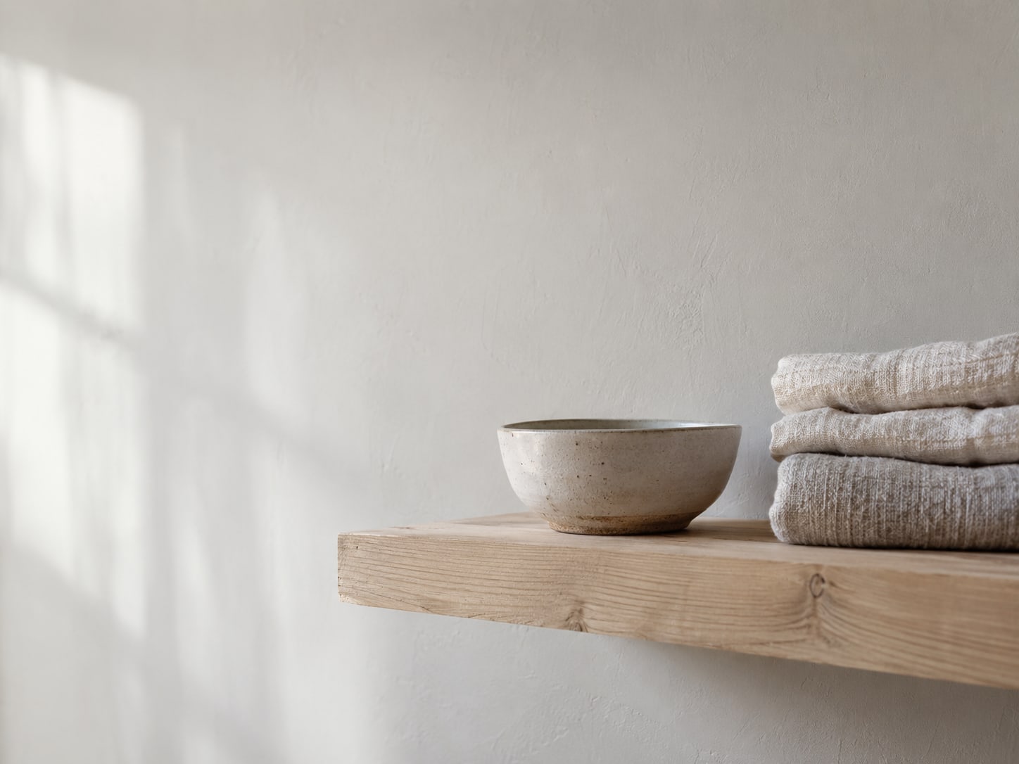 A wooden shelf holding a few small folded towels and a ceramic bowl in warm daylight.