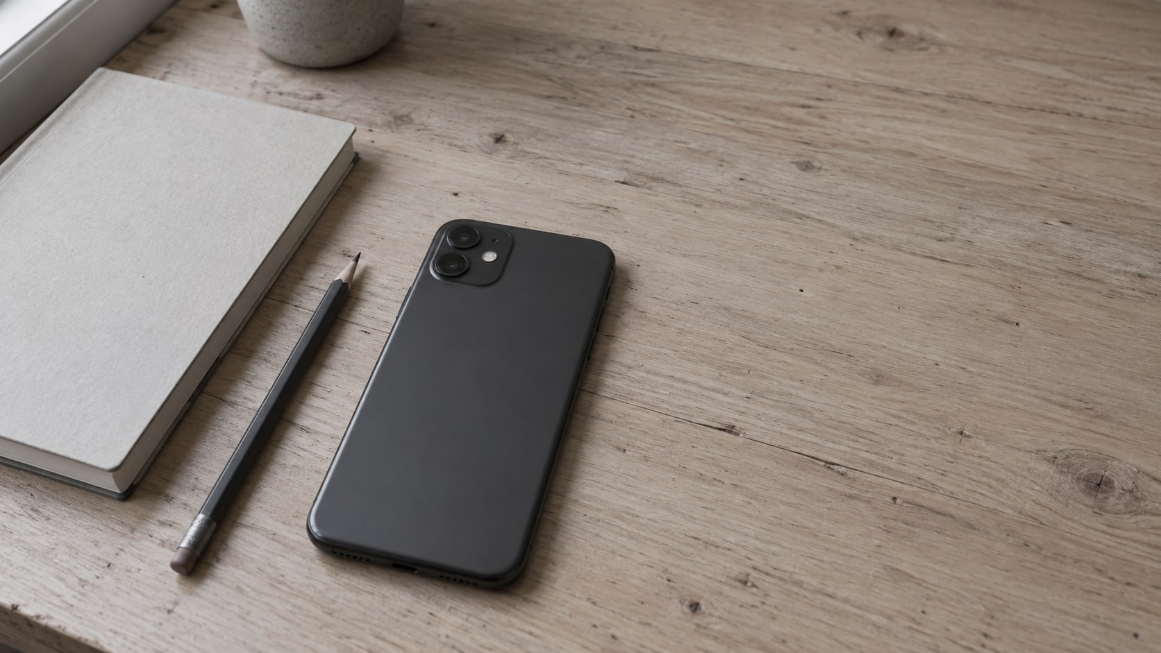 A smartphone lying face down on a light wooden desk beside a closed notebook and a pencil.