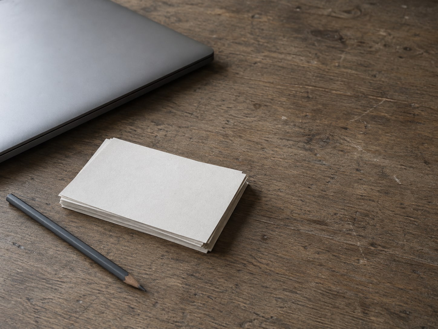 A closed laptop beside a small stack of blank index cards and a pencil on a worn wooden desk.