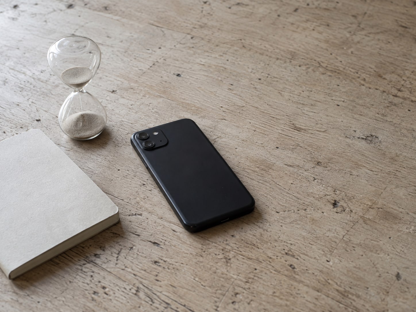 A phone lying face down on a wooden table next to a small hourglass and a closed notebook.