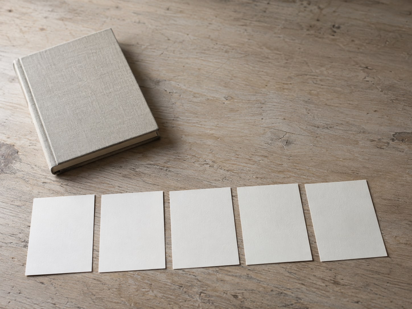 A closed notebook beside a row of blank index cards on a wooden desk, each card tucked slightly apart from the next.