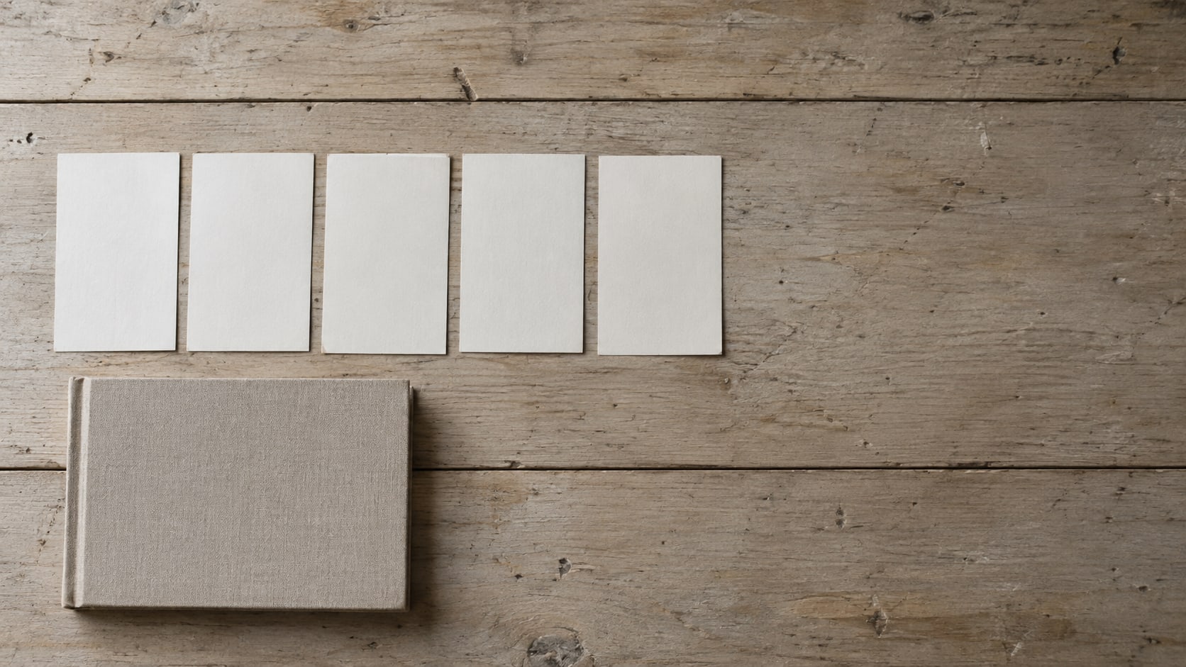 A row of blank index cards standing upright on a worn wooden desk beside a closed notebook.