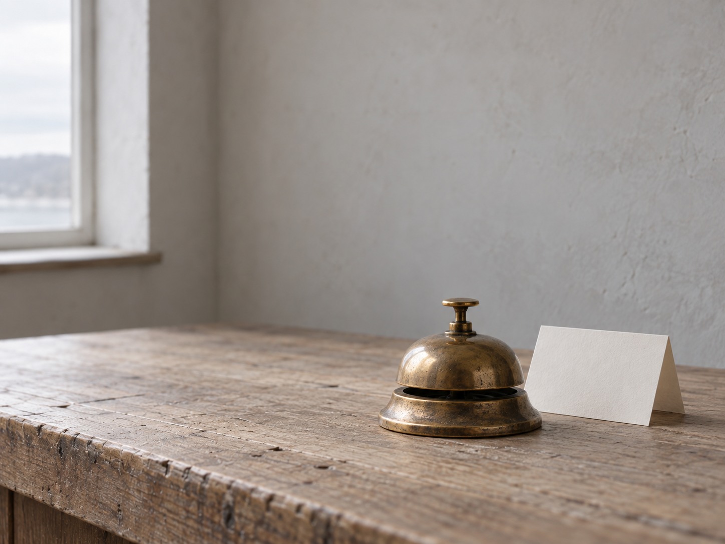 A small brass desk bell on a bare wooden counter beside a blank folded card.