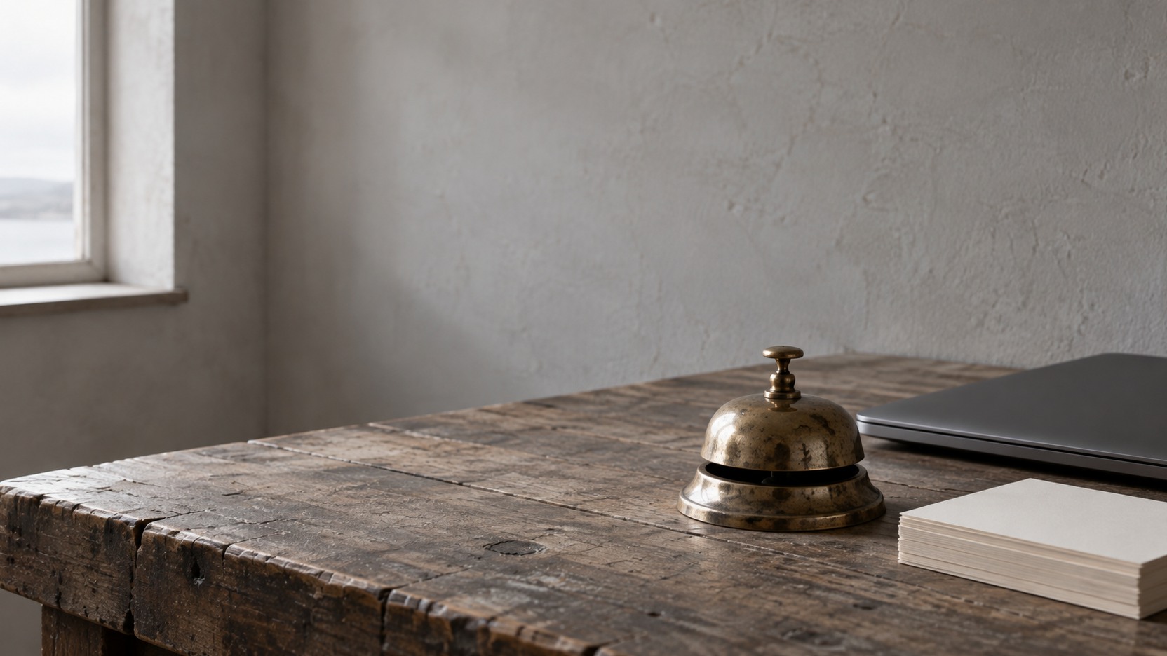 A small brass desk bell beside a closed laptop and a short stack of blank cards on a wooden counter.