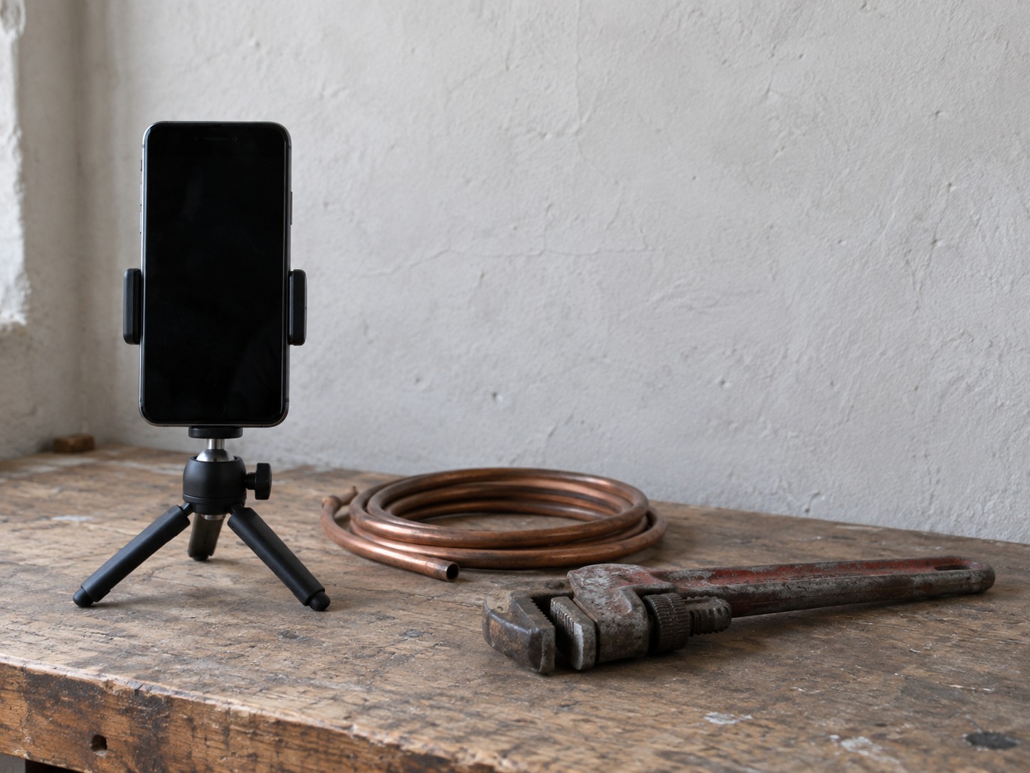 Un teléfono apagado sujeto a un trípode pequeño sobre la mesa de trabajo de un plomero junto a un rollo de tubo de cobre y una llave de tubo.
