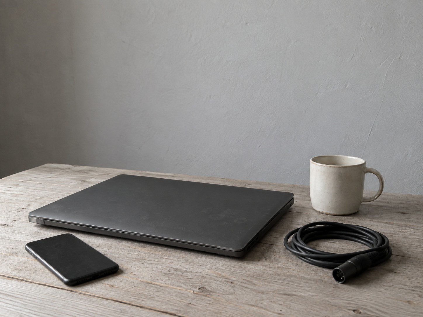 Una laptop cerrada, un teléfono boca abajo y un cable de micrófono enrollado sobre un escritorio de oficina junto a una taza de cerámica.