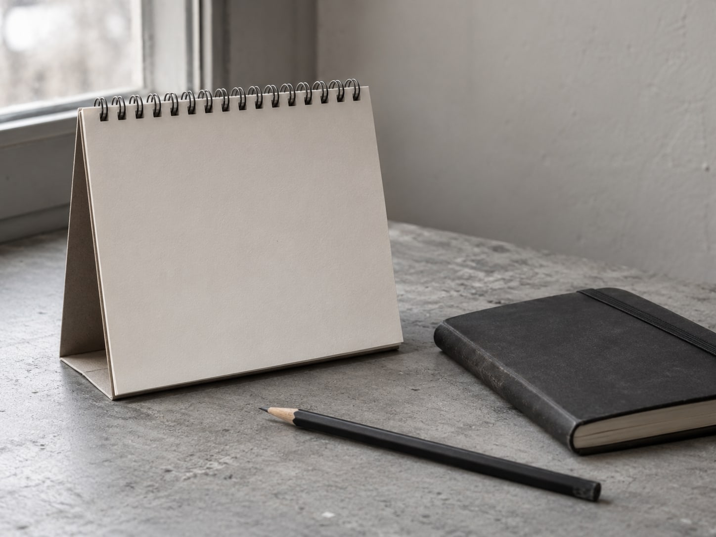 Un pequeño calendario de escritorio abierto en un mes en blanco junto a un cuaderno de bolsillo cerrado y un solo lápiz.