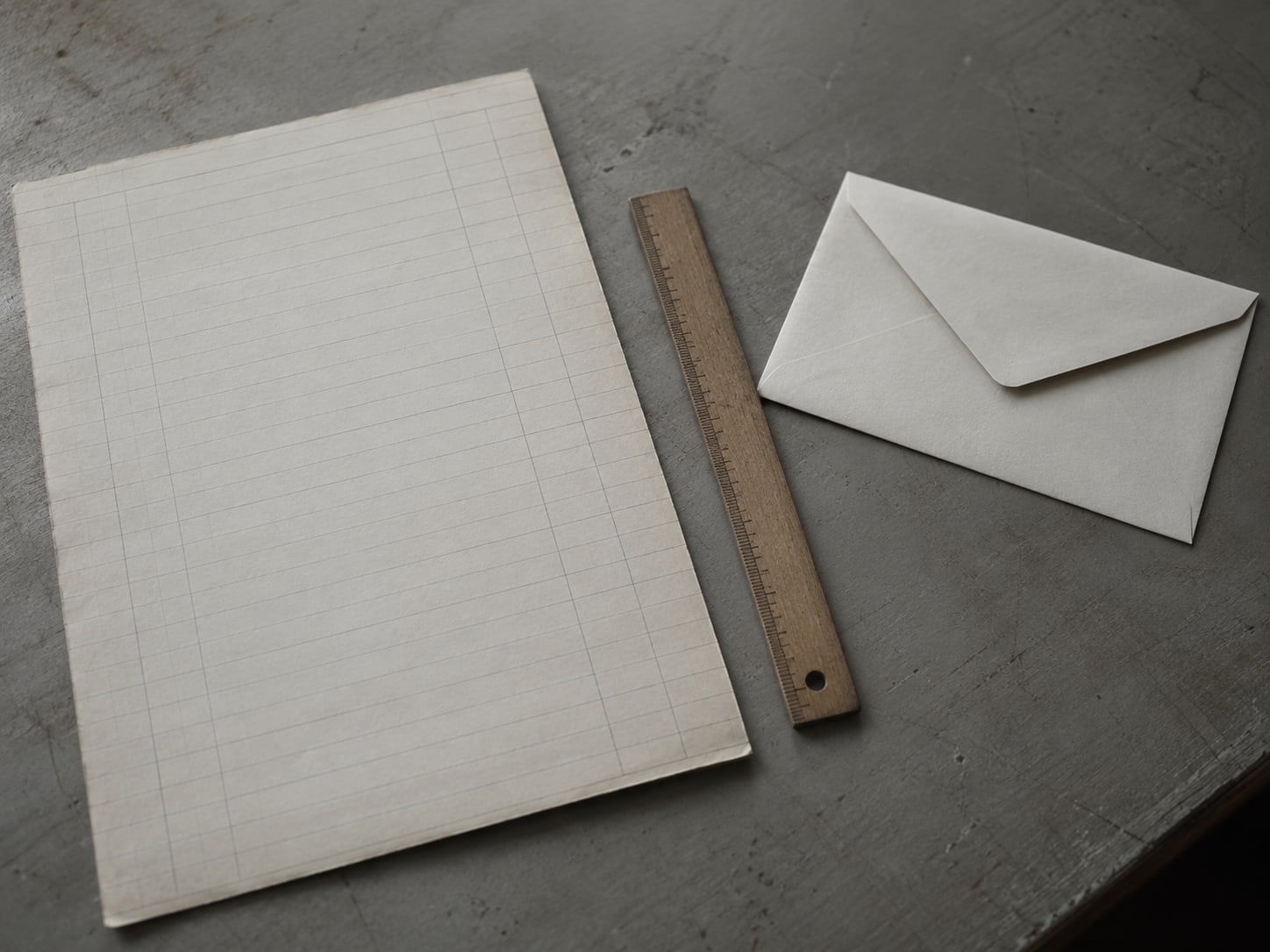 Una página de libro contable de papel con líneas tenues de lápiz junto a una regla de madera sencilla y un sobre sin abrir.