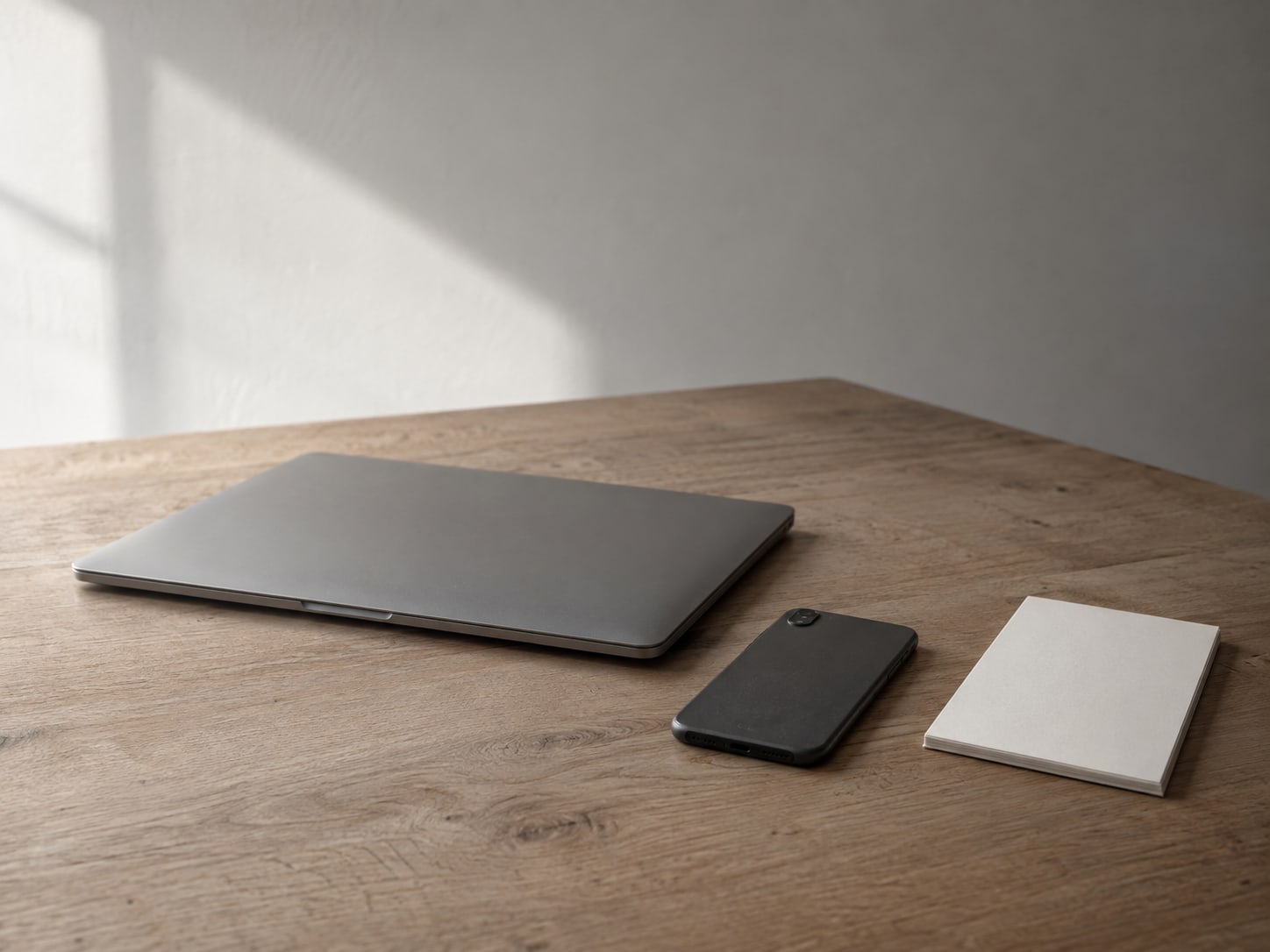 A closed laptop and a phone lying face down on a plain wooden desk beside a blank notepad.