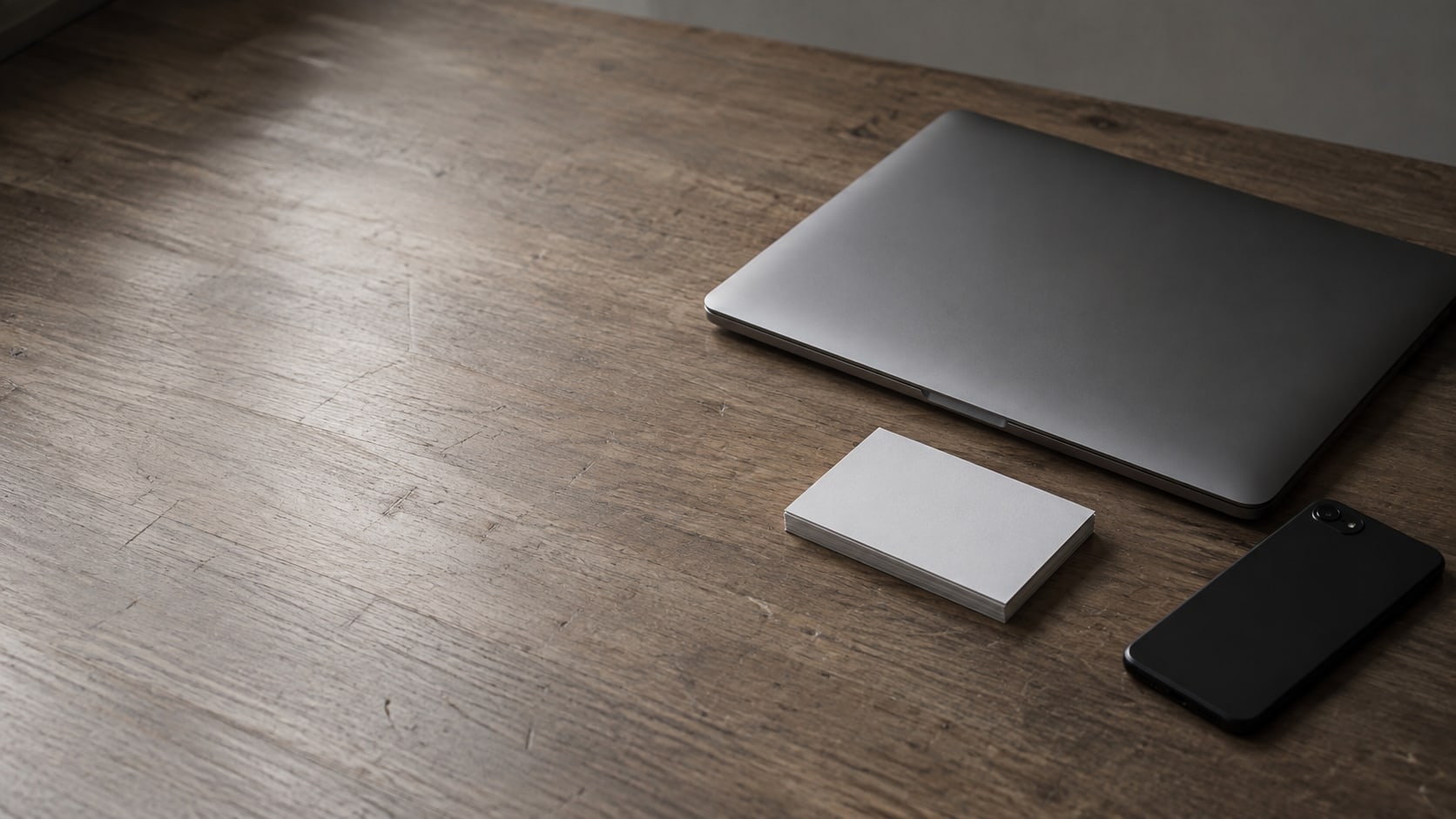 A closed laptop and a small stack of blank index cards beside a switched-off phone on a plain wooden desk.