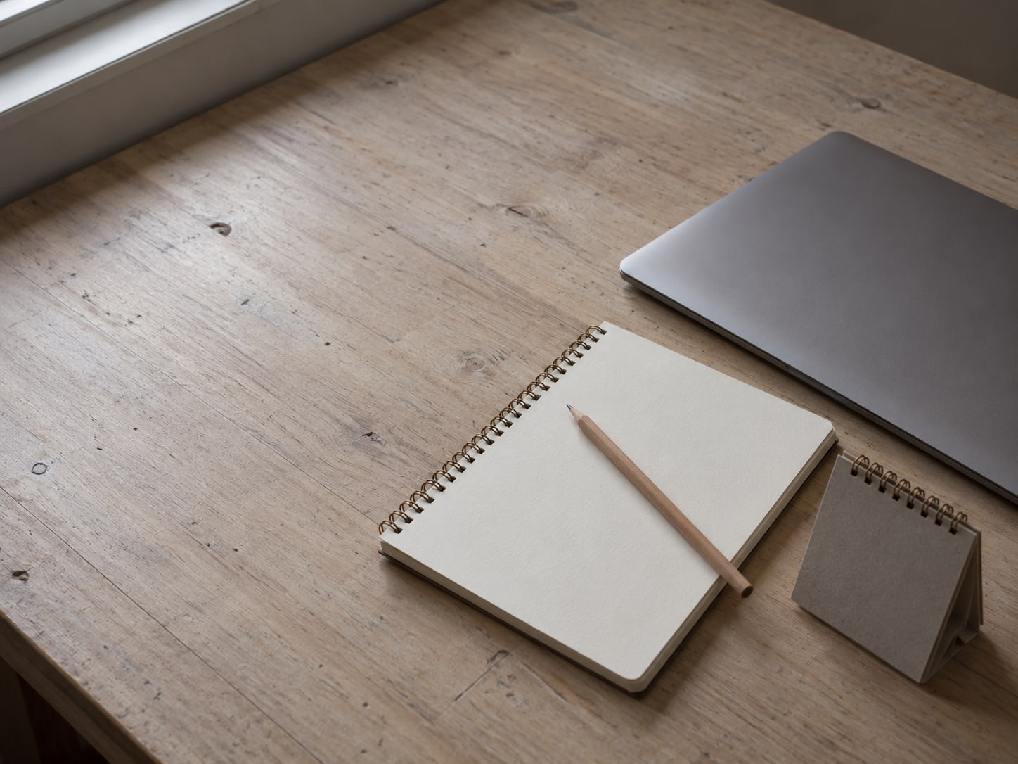 A pencil lying on a blank spiral notebook next to a closed laptop and a small desk calendar turned face down.