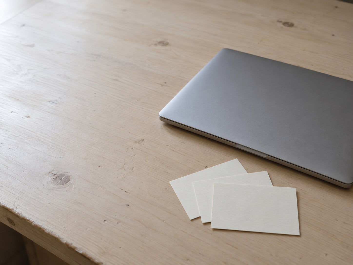 A closed laptop beside three blank index cards fanned out on a pale wooden desk.