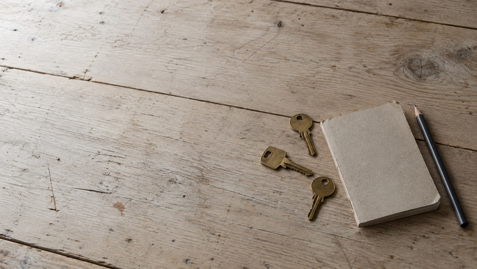 A notebook, a pencil and three plain brass keys laid out on a wooden desk.