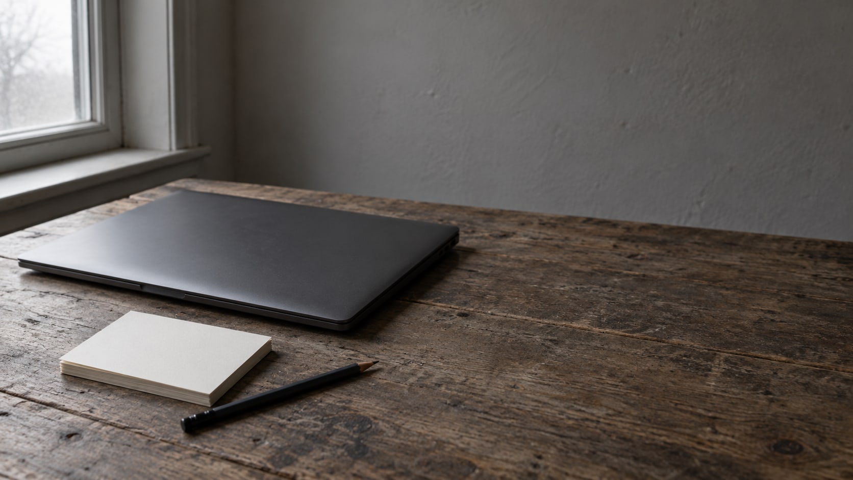 Una laptop cerrada y una pequeña pila de fichas en blanco sobre un escritorio de madera gastada, con un lápiz al lado.