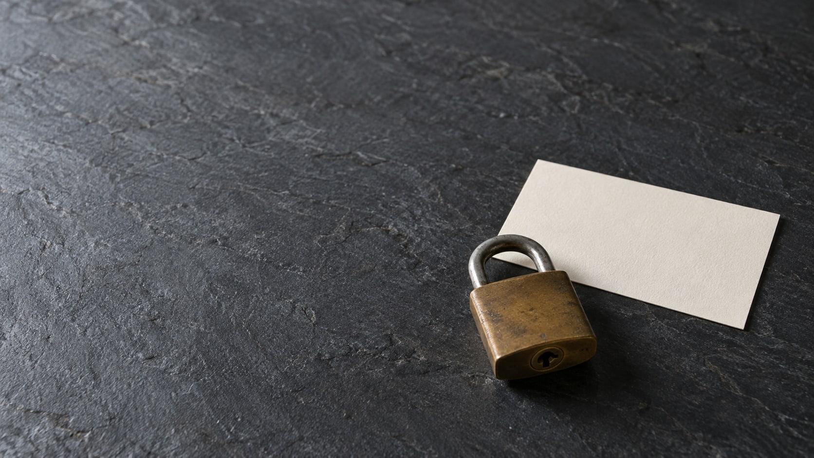 A small brass padlock and a blank white card on a dark slate surface, lit softly from one side.