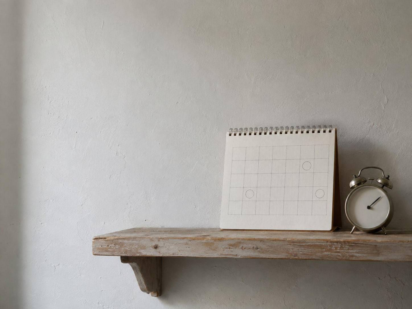 A wall calendar with pencil marks on several dates next to a small alarm clock on a wooden shelf.