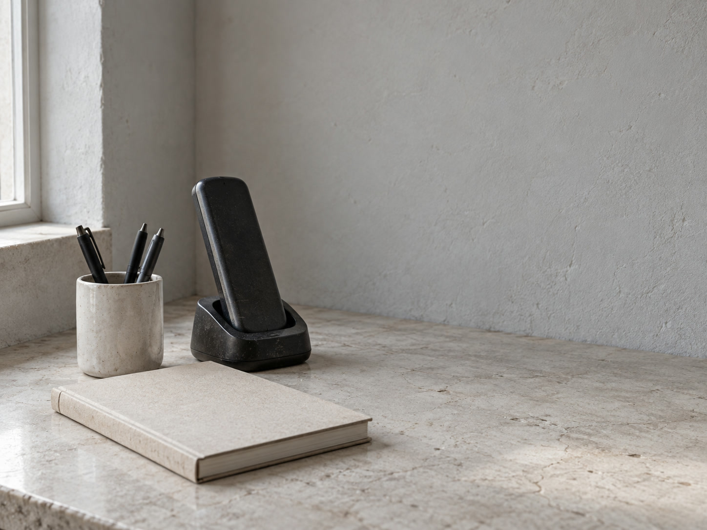 A cordless desk phone resting beside a closed paper appointment book and a small ceramic cup of pens on a white counter.