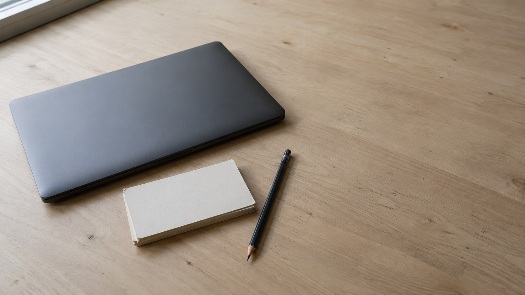 A closed laptop, a small stack of blank index cards and a pencil resting on a clean wooden desk.