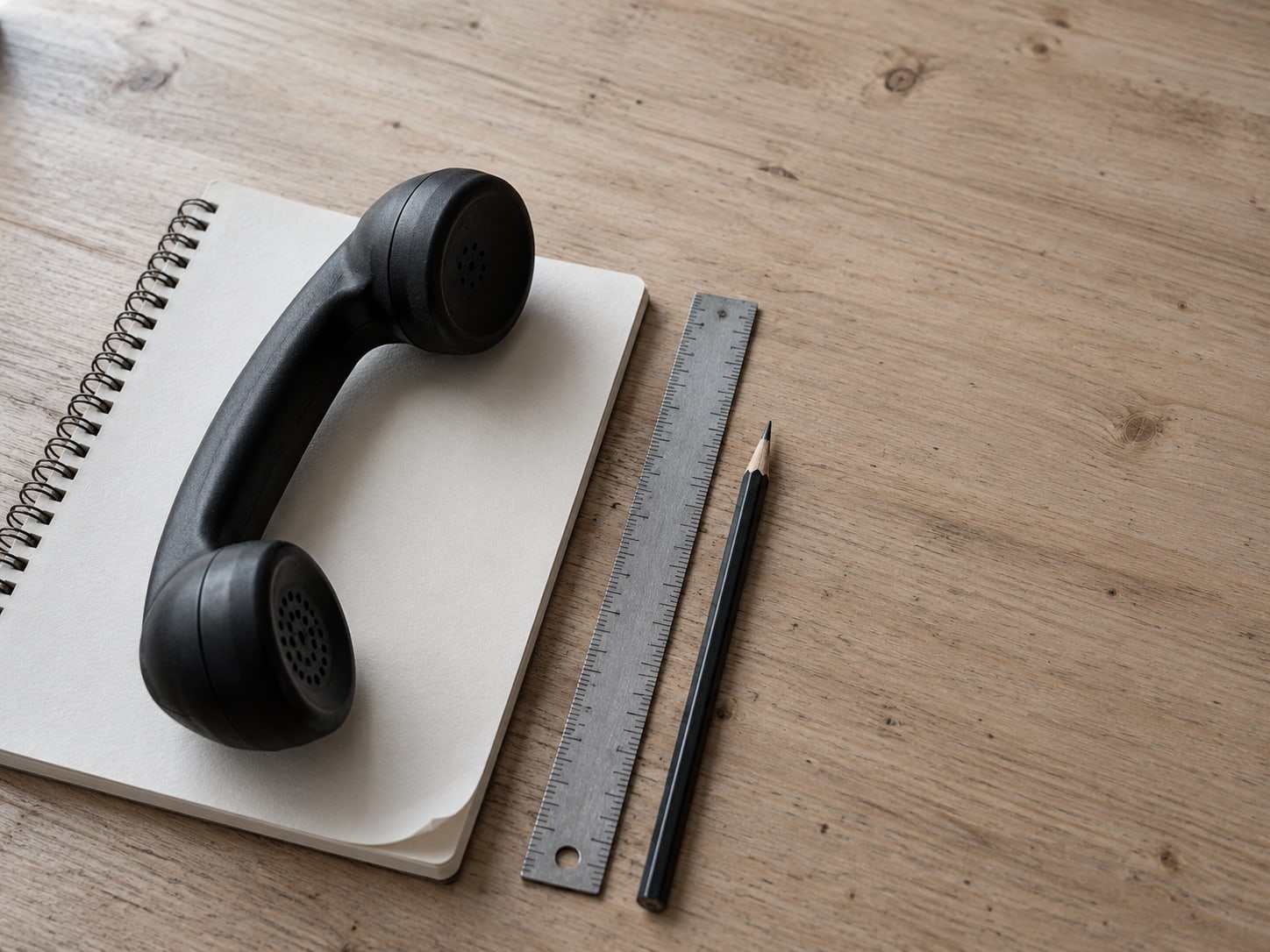 Un auricular de teléfono sobre una libreta en blanco, junto a una regla y un lápiz afilado sobre una superficie de madera.