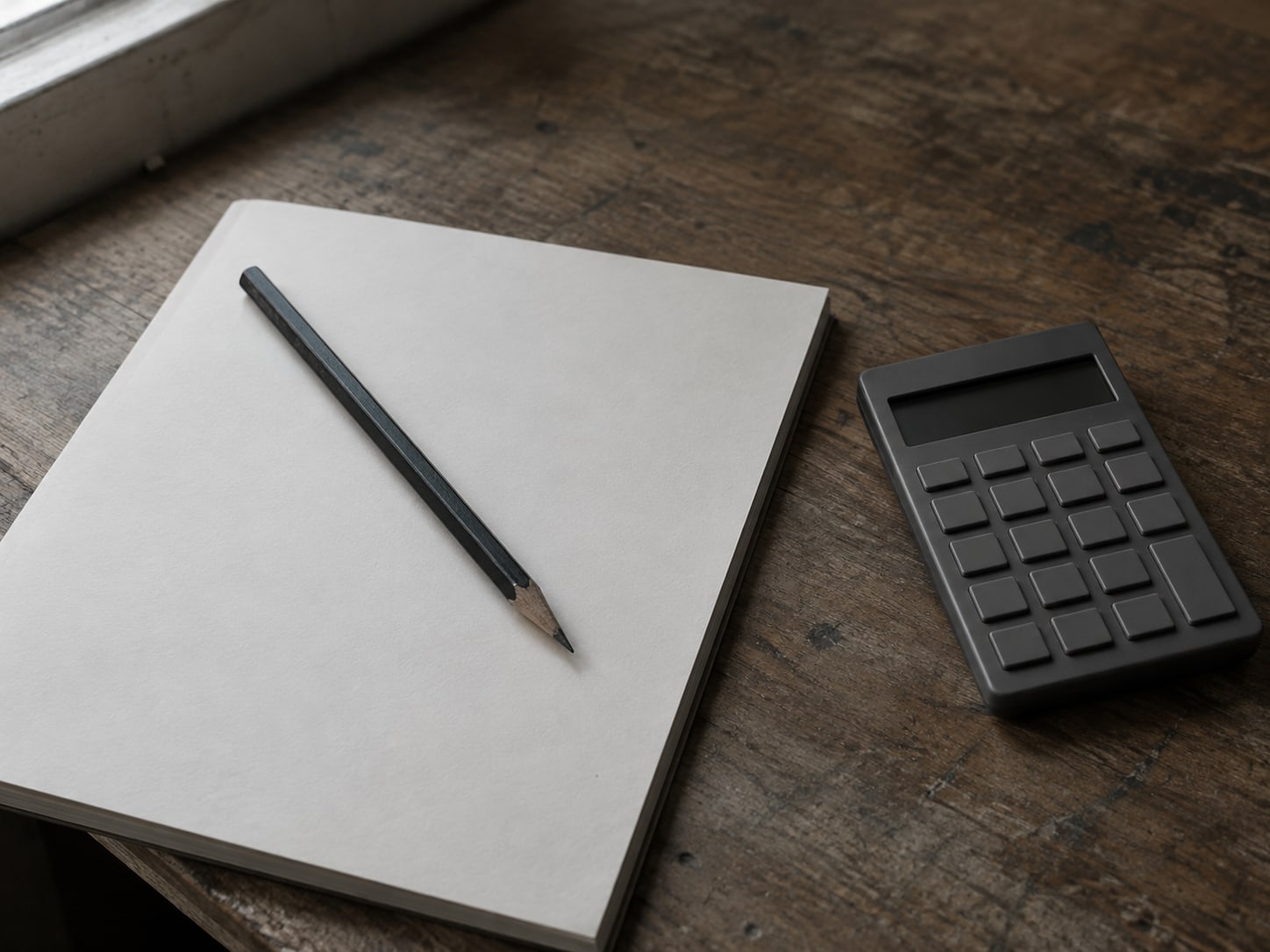 A pencil lying across a blank ledger page next to a small calculator on a desk.