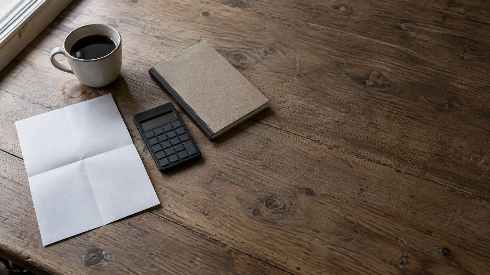 A folded blank invoice and a small calculator on a wooden desk beside a closed notebook and a cup of coffee.