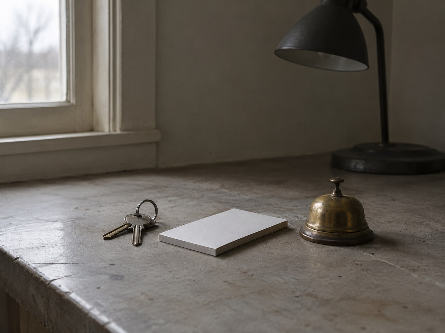 A pair of keys, a blank notepad and a small brass bell resting on a worn counter beside an unlit desk lamp.